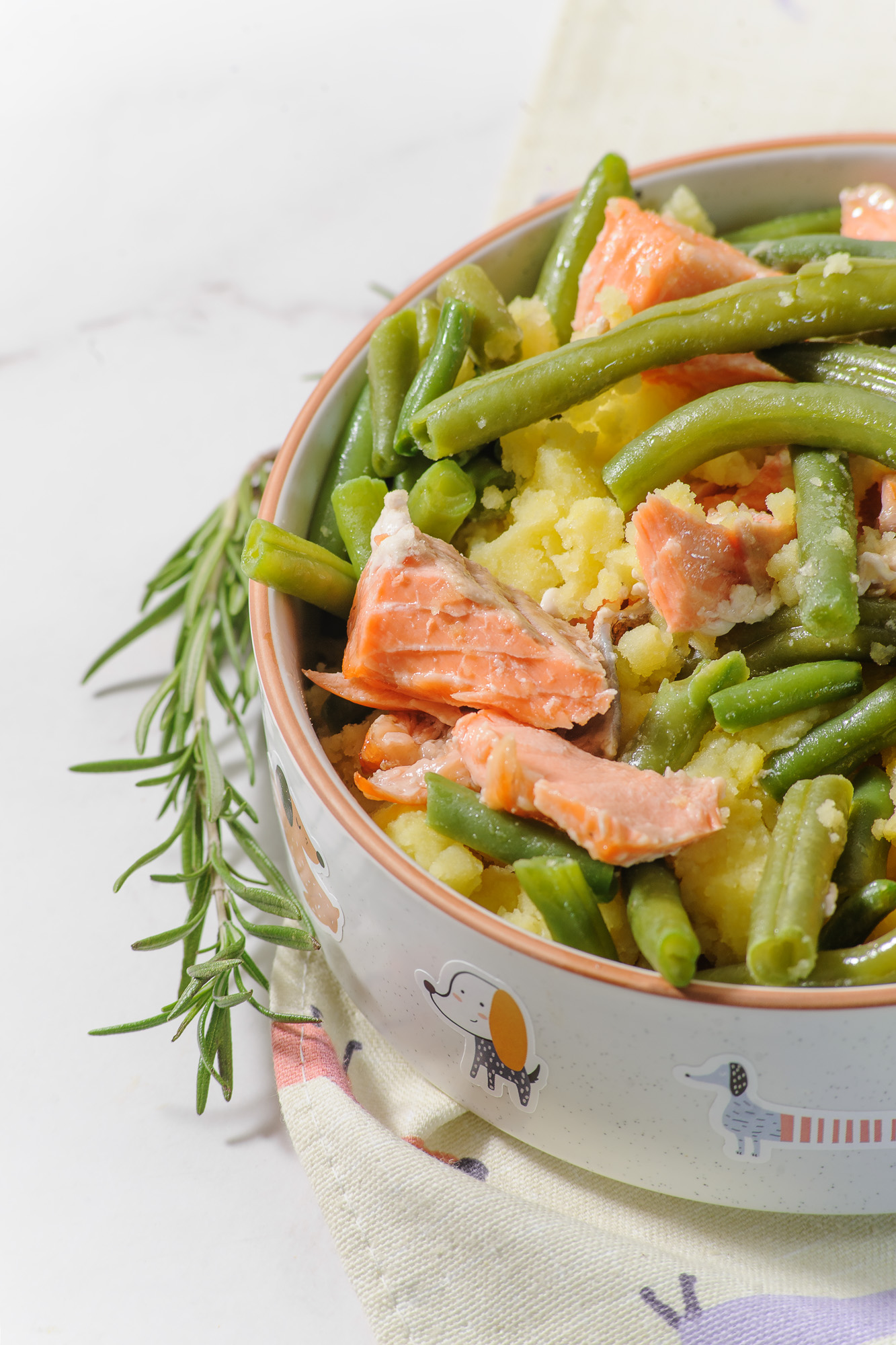Baked salmon bits mixed in a bowel with green beans and sprig of rosemary on the side of the bowl.