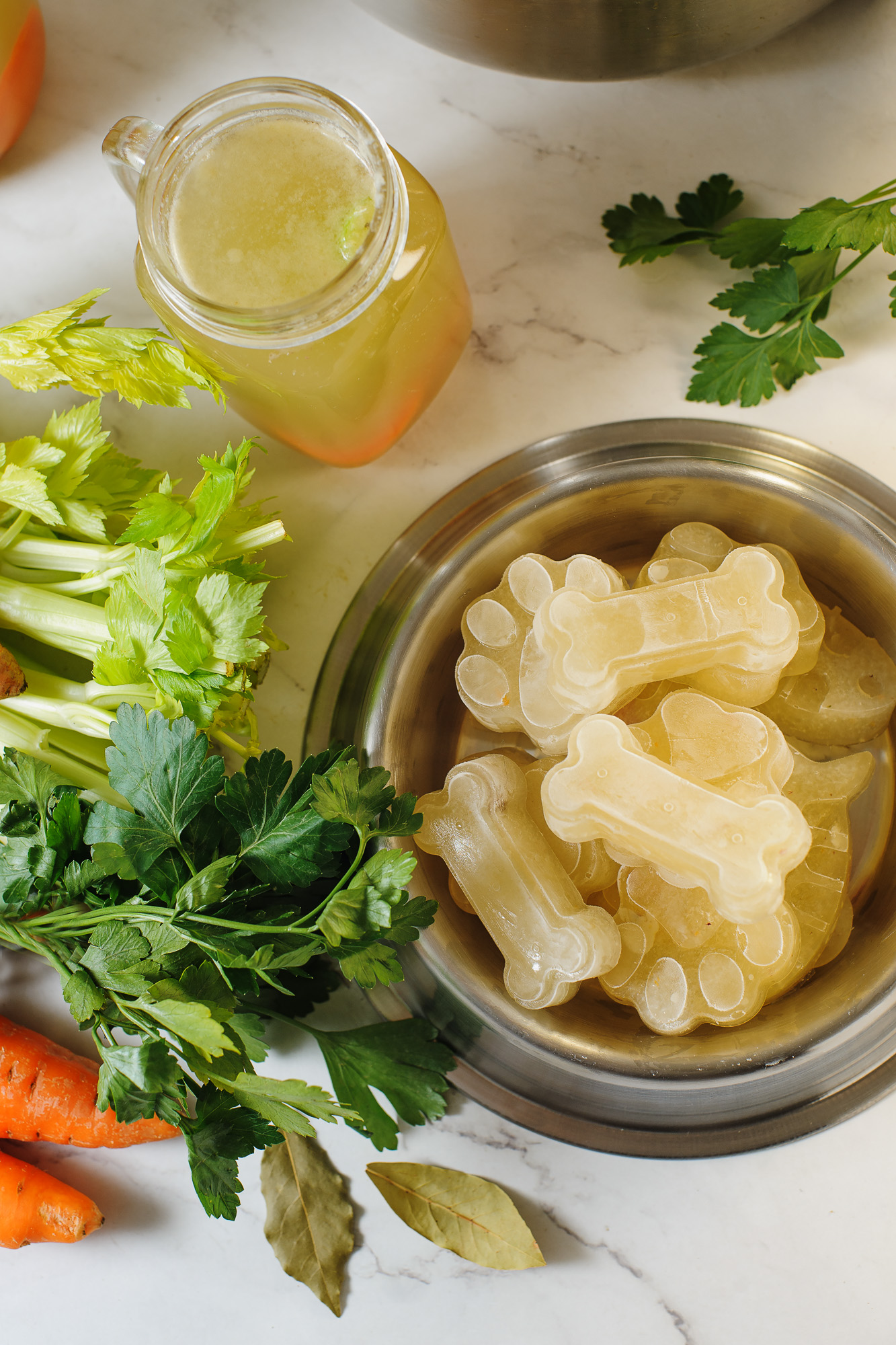 A photo of frozen bone broth in the shapes of dog treats and dog paws in a silver bowl. There is celery and a jar of bone broth on the counter. 