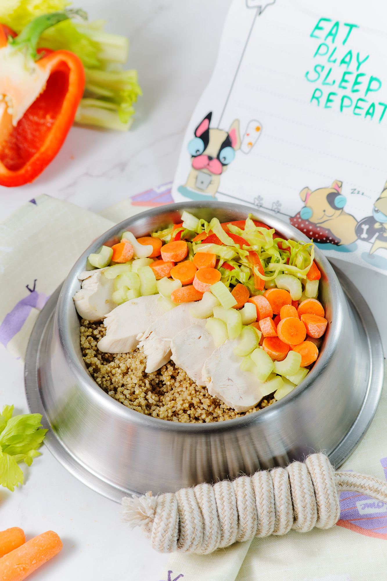 A bowl is filled with chicken, carrots, celery and quinoa with peppers and a dog treat in the background