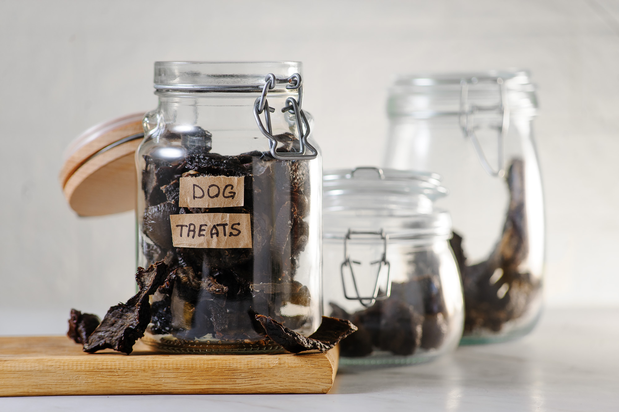 Dehydrated Liver, Kidneys and Heart dog treats in three seperate jars with open glass lids. One jar sits on a wooden cutting board with treats in front on the cutting board. 