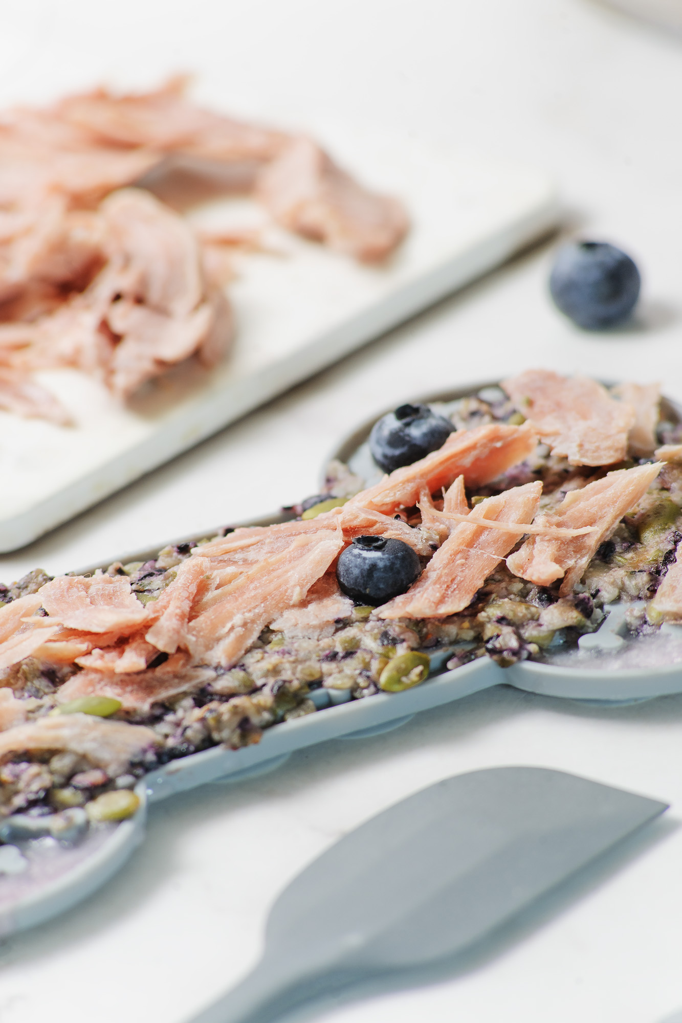 Turkey meat shredded on top of a lick mat for dogs in the shape of a bone with two blueberries on top. There is shredded blueberries on a white cutting board in the background. 