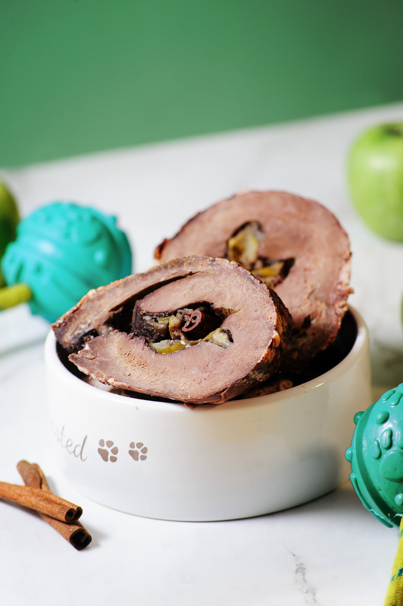 A bowl with a beef heart roasted and cut in half with blue dog toys and a green apple with a green background. 