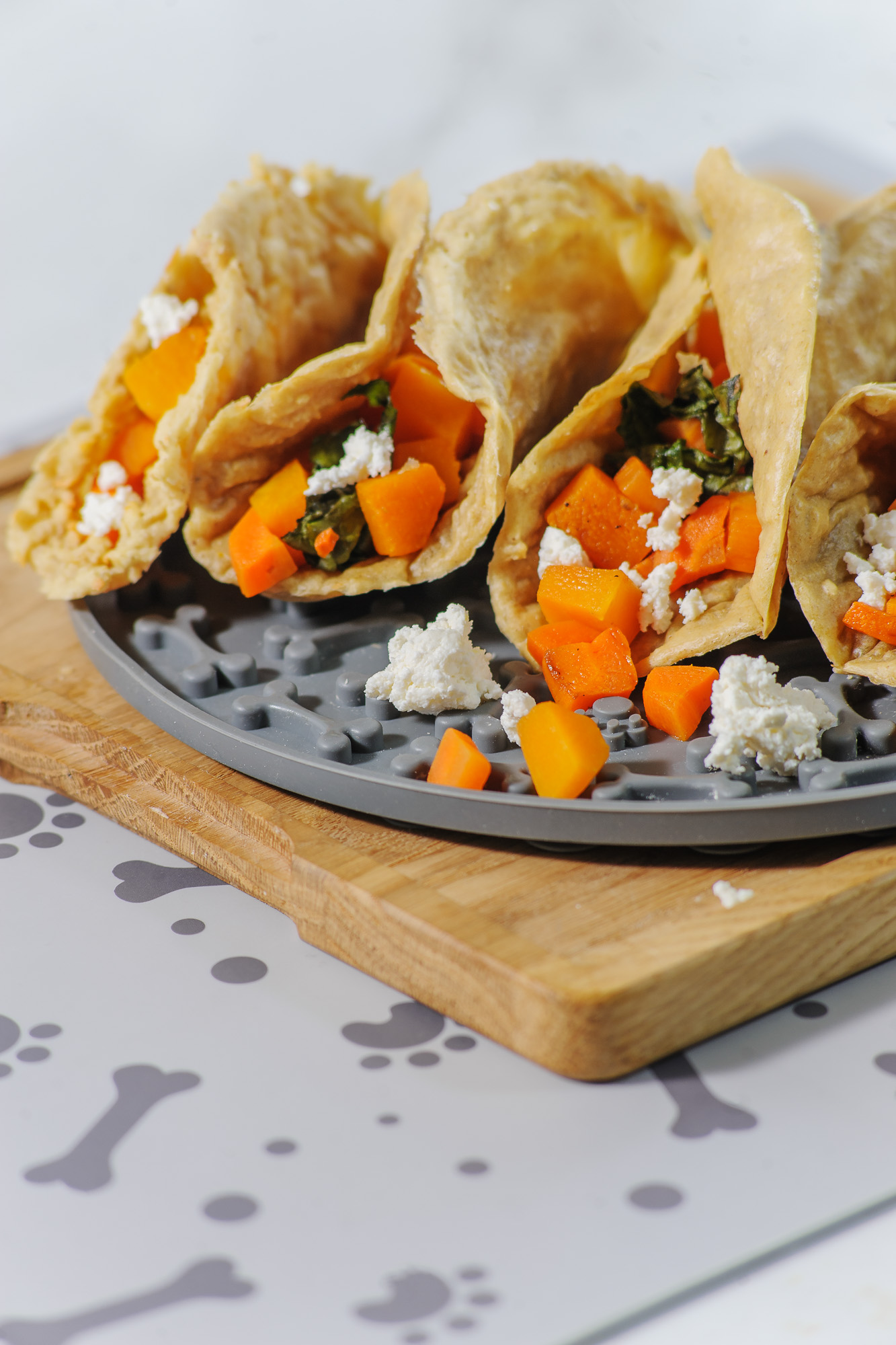 Tacos on a lick mat for dogs that is placed on a cutting board. The tacos have orange yams and cheese coming out of them. 